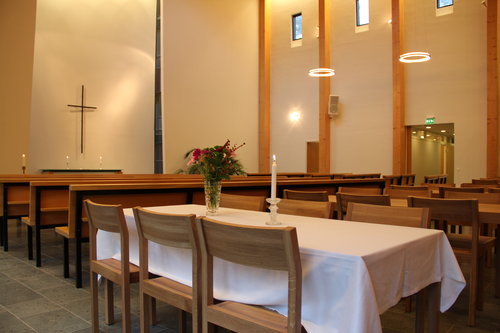 Laajakosken kappelin kahvitustilat, tuoleja ja pöytä, jolla valkoinen liina ja kynttilä. Taustalla penkkirivit ja alttari.