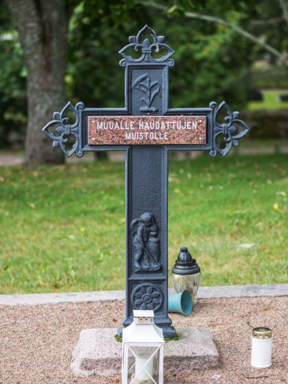 Risti hiekkamaalla, taustalla kasvillisuutta, ristin juurella lyhtyjä ja kynttilöitä.
