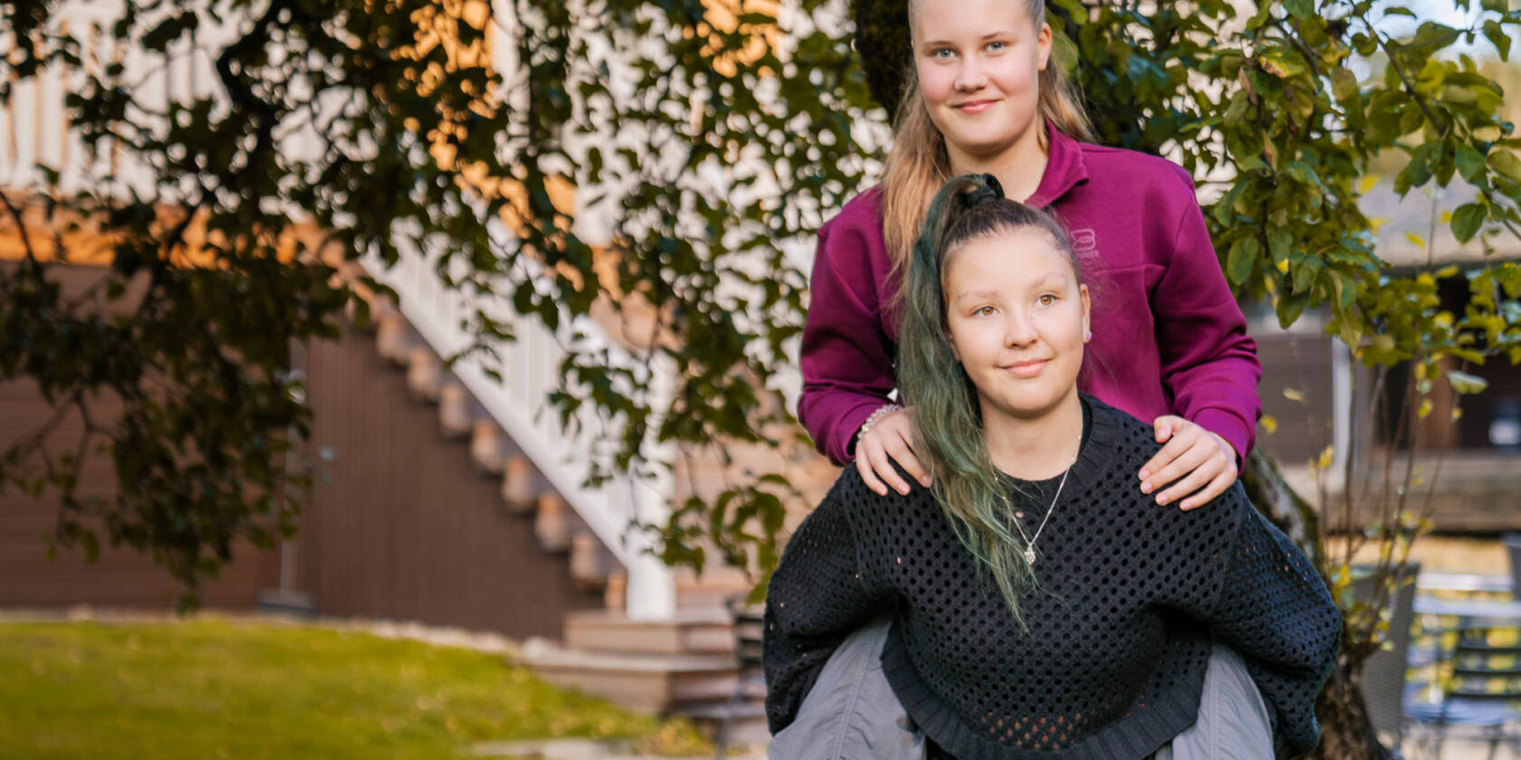 Kaksi nuorta ulkona puun alla, toinen on toisen reppuselässä.